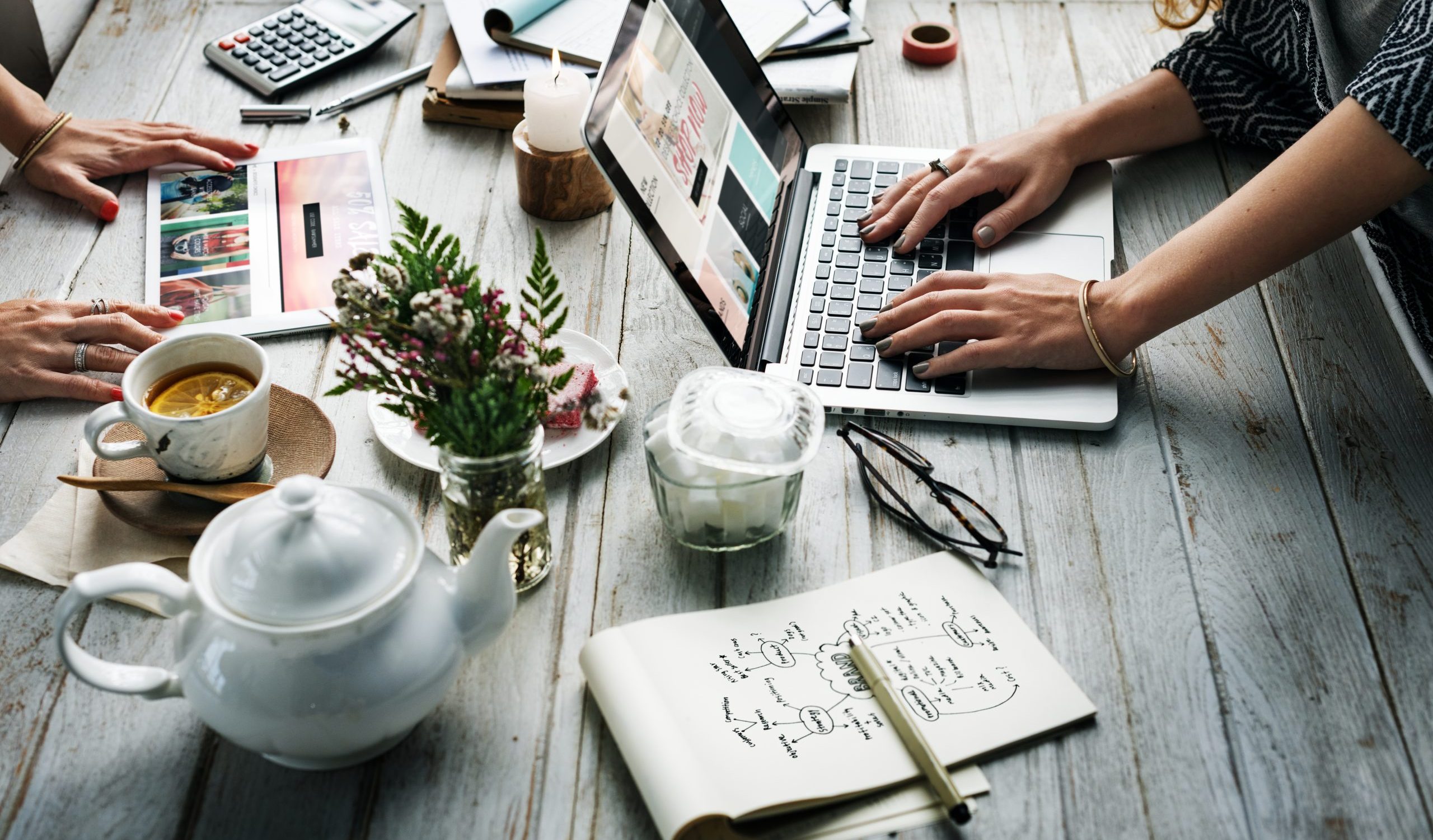 Zwei Hände, die an einem Schreibrisch am Laptop tippen. Auf dem Tisch stehen Kaffee und es wird an einem Marketing Plan gearbeitet